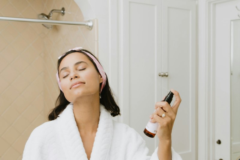 A woman in a robe spraying facial mist in a bathroom mirror